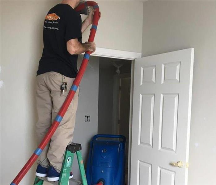 male technician in black t-shirt with SERVPRO logo and khaki pants standing on ladder inserting duct cleaning hose in vent