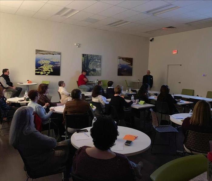 16 people in room sitting around round tables listening to restoration teacher