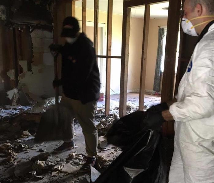 two men wearing face masks clean up pile of fire damaged debris in room with studs
