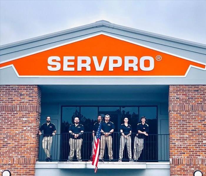 five guys and one woman in black SERVPRO shirt and khaki pants standing on balcony with American flag and SERVPRO sign