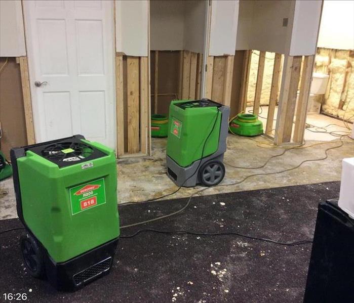 green dehumidifiers and fans in empty room with sheetrock removed and studs exposed from the floor to four feet up the walls